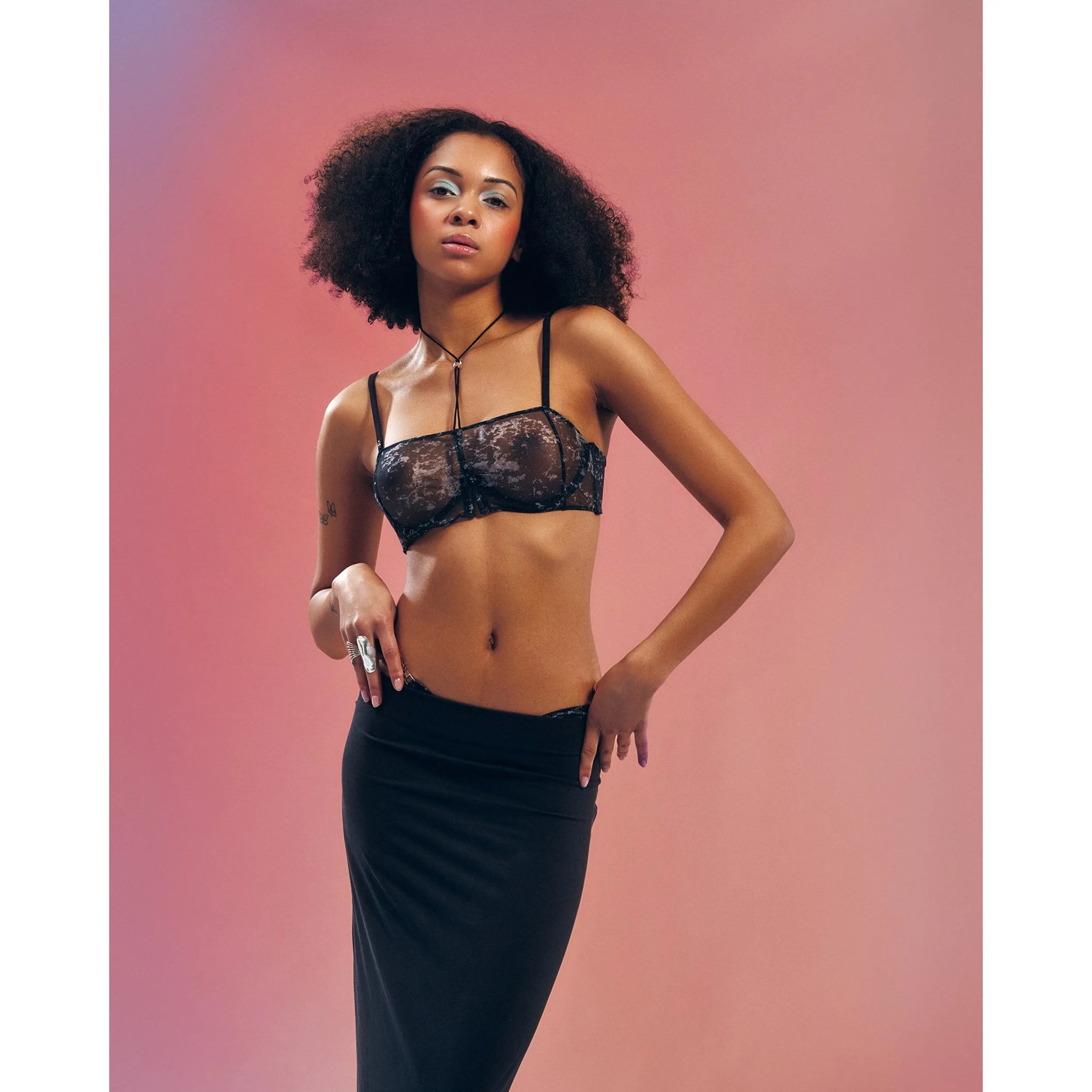 Soutien-gorge Corrosis noir transparent froncé, armatures réglables, motif unique, porté par un mannequin sur fond rose.