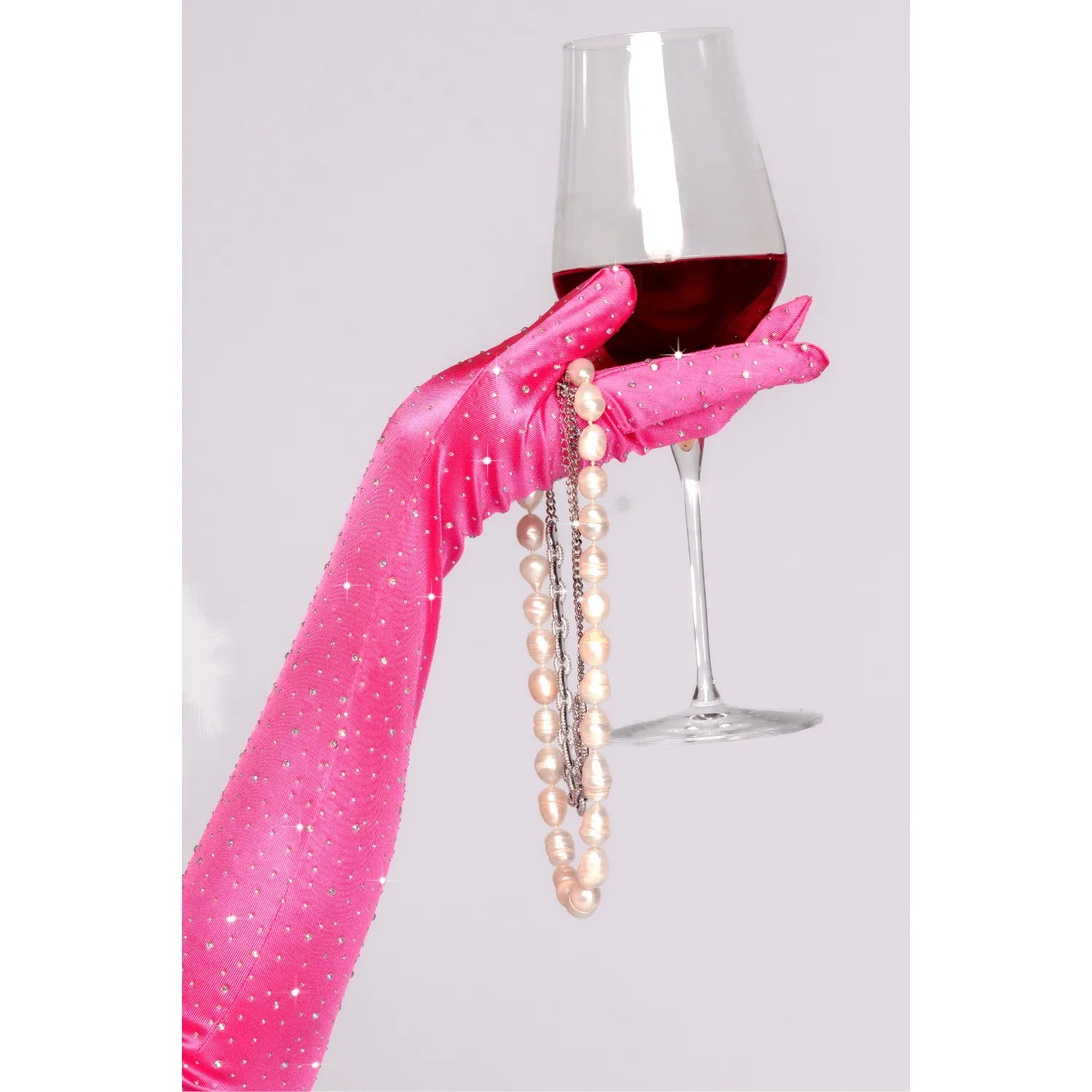 Gants satinés roses extensibles avec strass, élégants, tenant un verre de vin et un collier de perles.