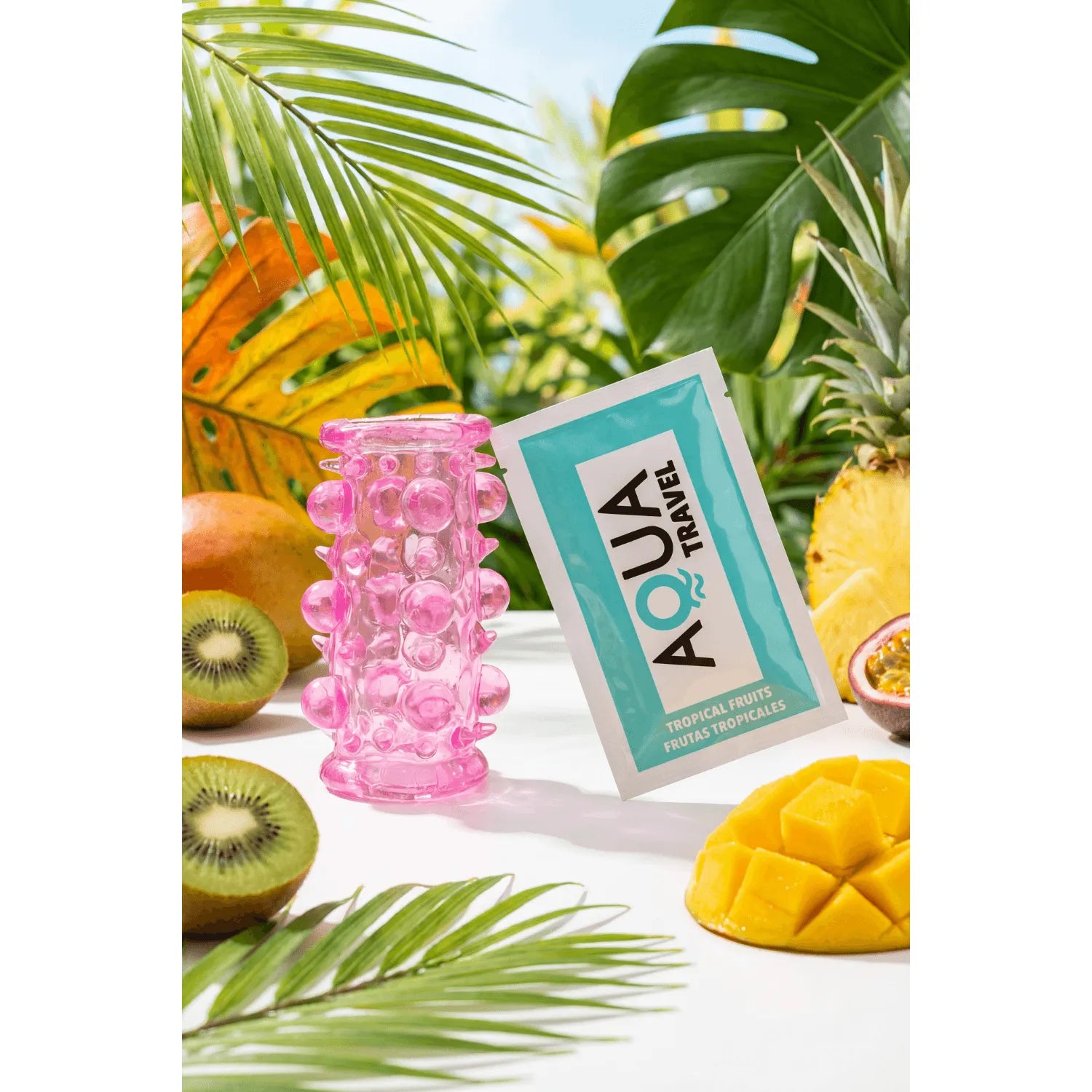 Gaine pénienne rose à picots et boules en relief, ultra-extensible, posée sur une table avec fruits tropicaux.
