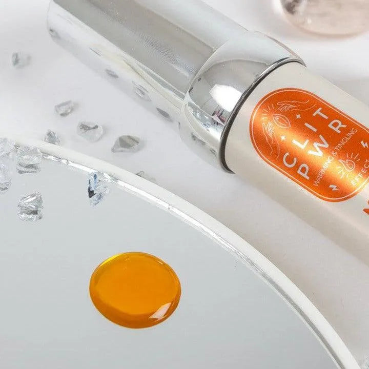 Baume clitoridien stimulant mangue, flacon argenté avec étiquette orange, goutte de baume sur miroir, cristaux décoratifs.