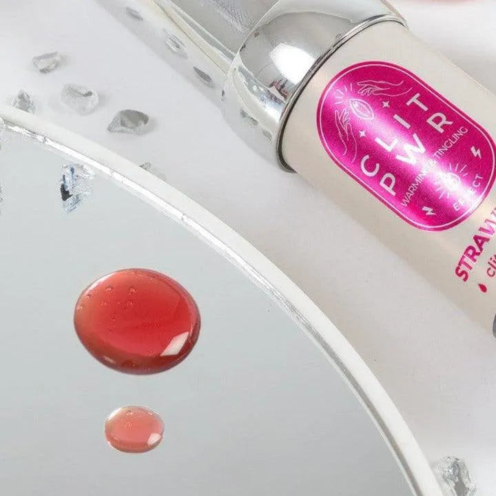 Baume stimulant clitoris fraise, tube blanc et rose, effet vibrant et chauffant, sur fond miroir avec gouttes rouges.