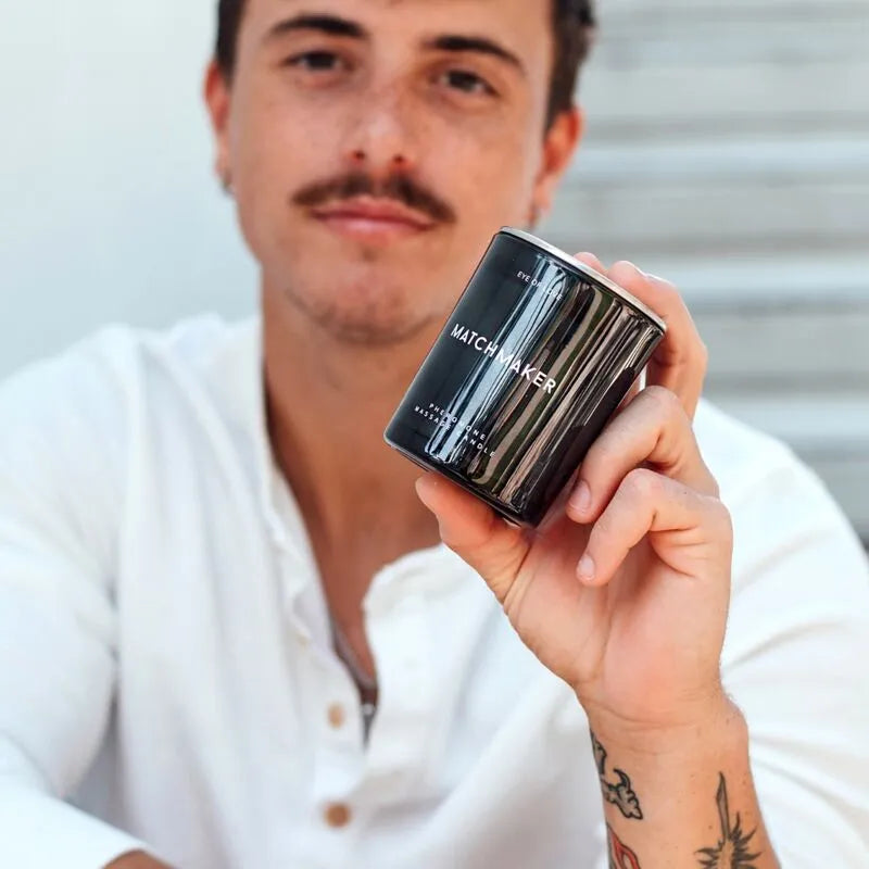 Bougie noire de massage aux phéromones, tenue par une personne, avec étiquette blanche, fond flou.