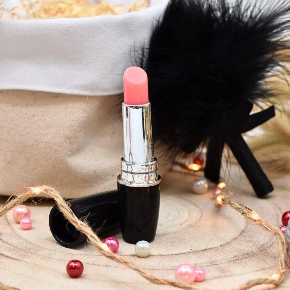Box cadeau bien-être naturelle avec rouge à lèvres rose, perles colorées et plumes noires sur fond en bois.