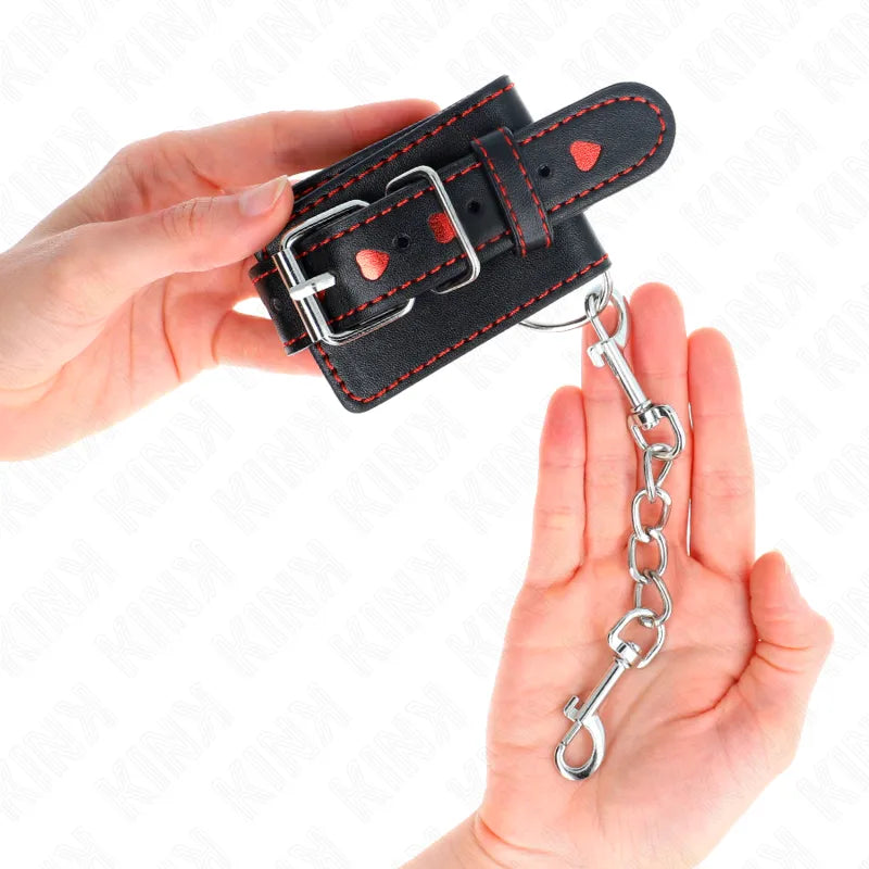 Bracelet BDSM en cuir noir ajustable avec coutures rouges, chaîne métallique et boucle, tenu par deux mains.