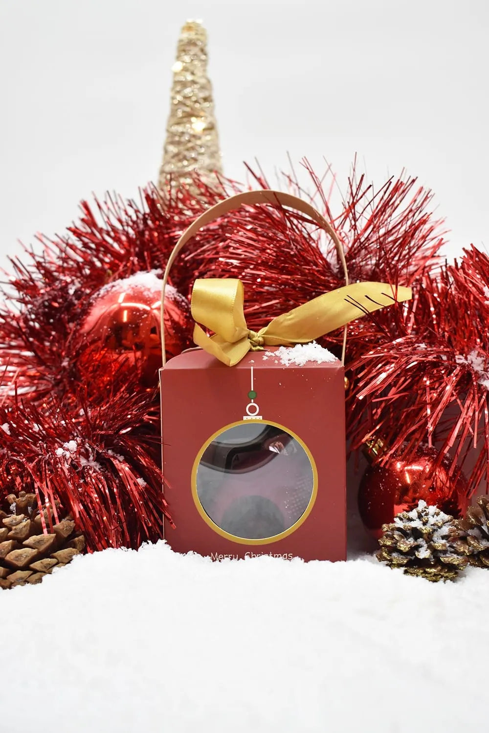 Coffret Coquin Noël rouge avec ruban doré, entouré de décorations festives rouges et or, sur fond de neige.