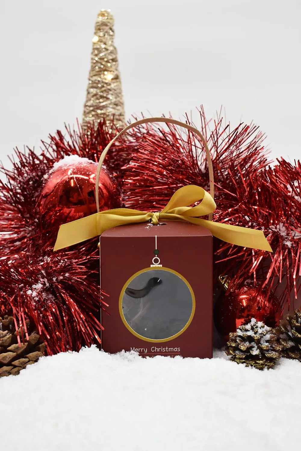 Coffret cadeau chocolat Noël rouge avec ruban doré, entouré de décorations festives rouges et dorées sur fond enneigé.