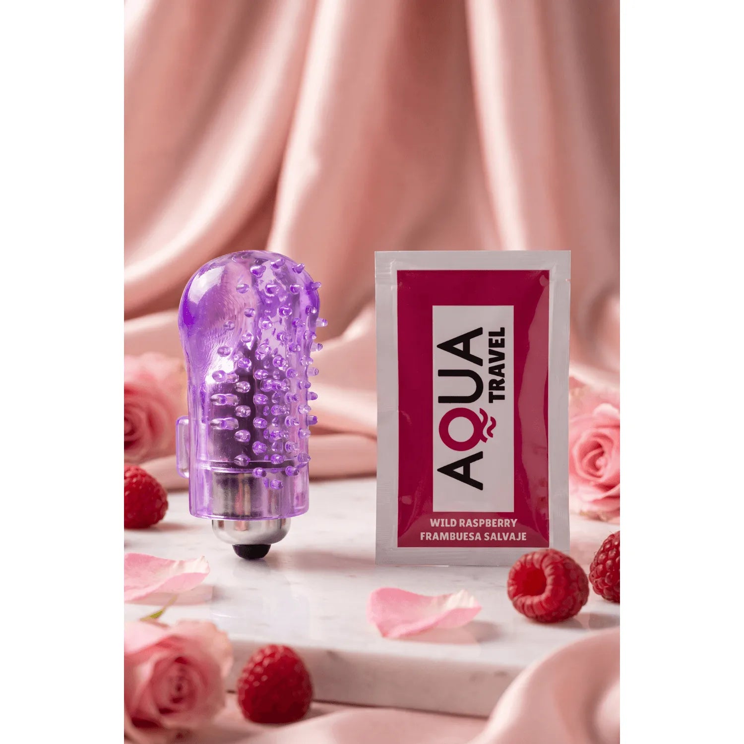 Doigt vibrant violet texturé avec picots et dosette de lubrifiant Aqua Travel, posé sur fond rose avec framboises.